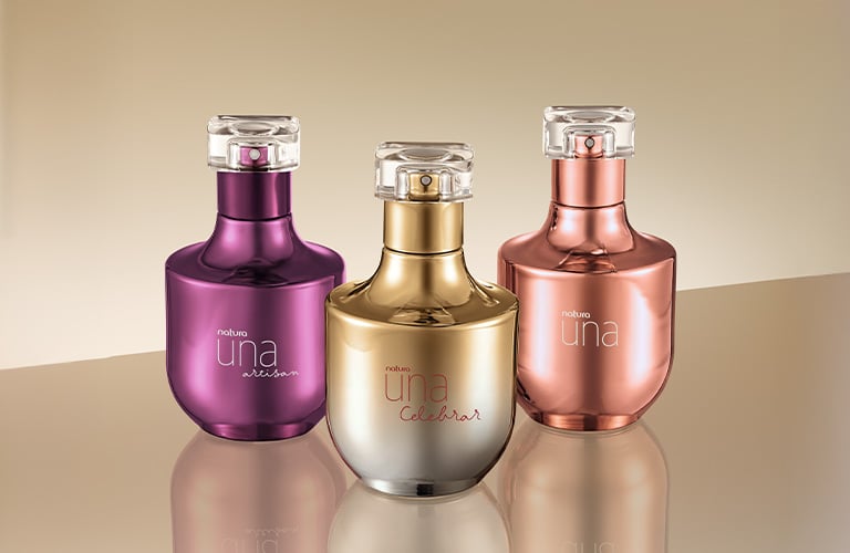 Foto de tres frascos de perfume Una de Natura, en colores morado, dorado y cobre, con reflejo en la superficie, sobre un fondo beige.
